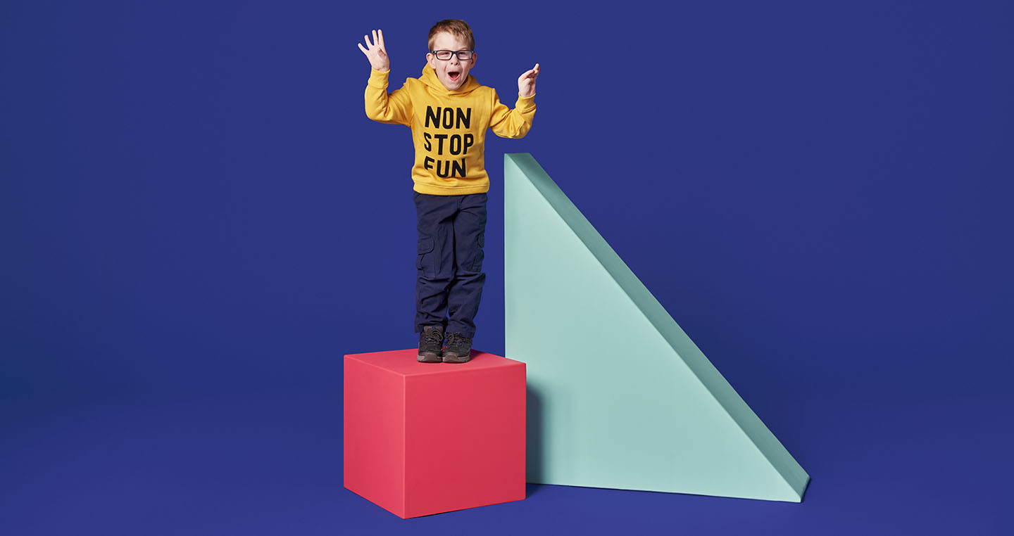 Child standing on a bright pink cube next to a large blue triangle against a purple background.