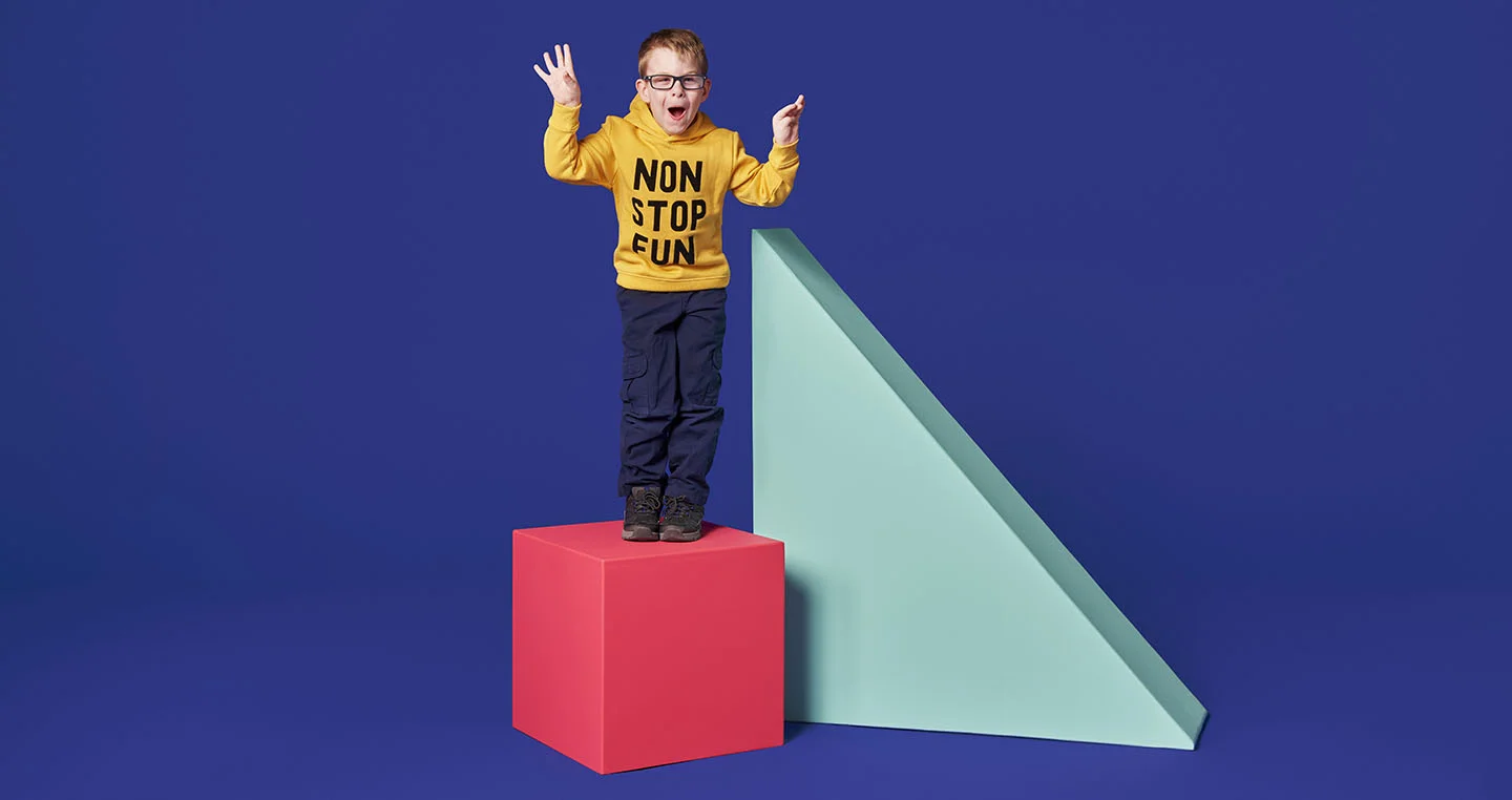 Child standing on a bright pink cube next to a large blue triangle against a purple background.