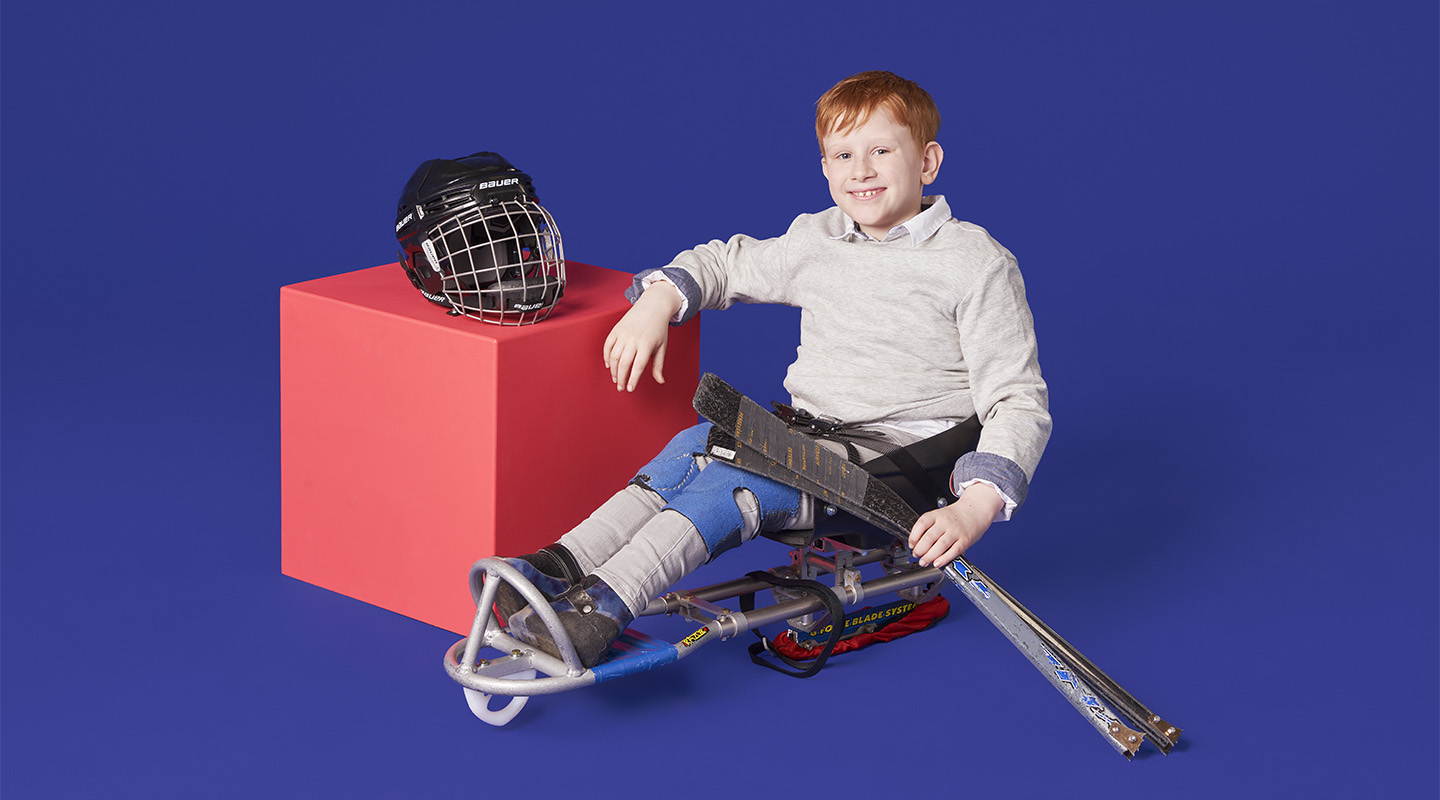 Still of a young sledge hockey player leaning against a vibrant pink cube on a purple background.