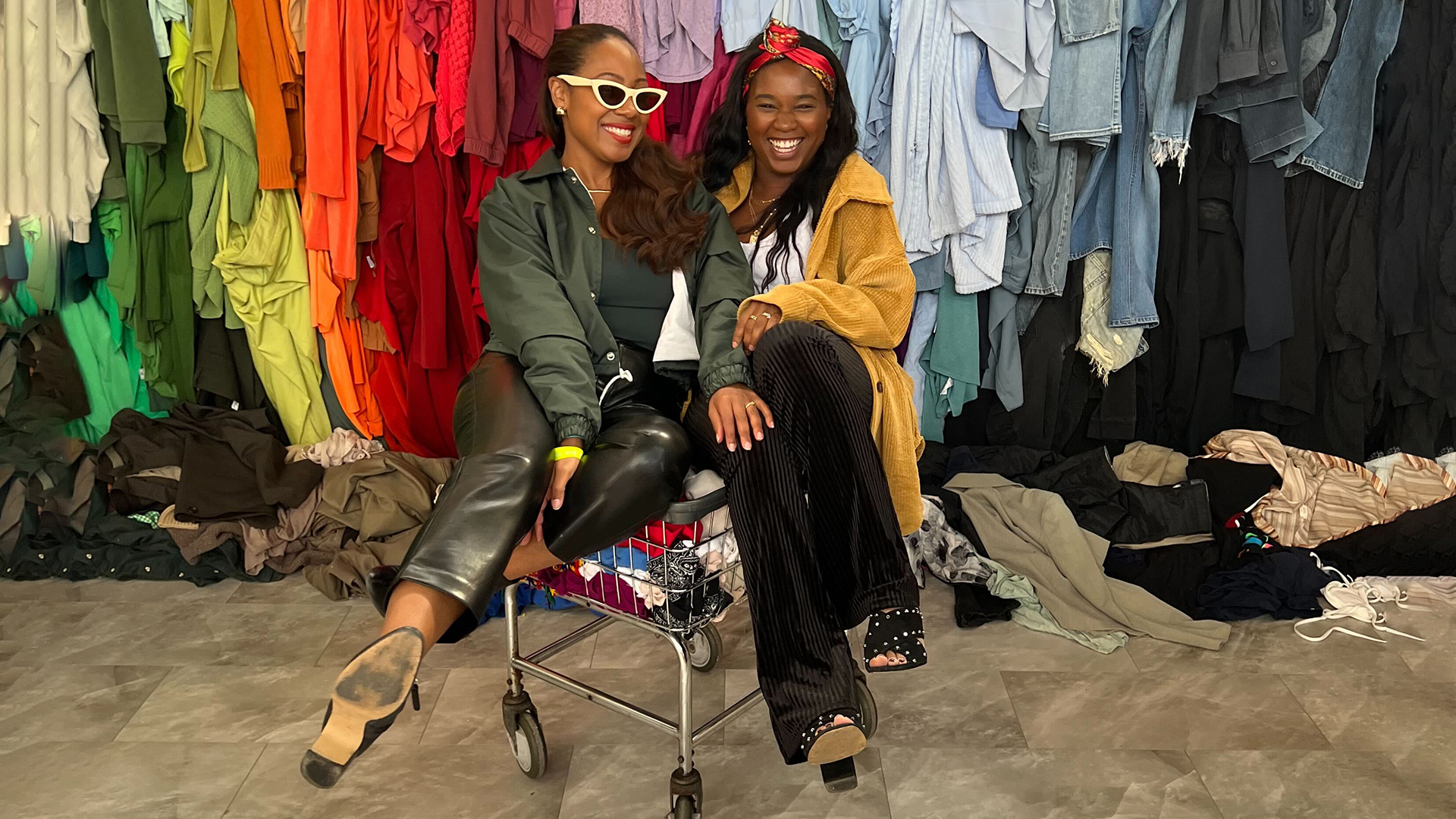 UpHouse team members Erica and Sade posing in a shopping cart in front of piles of clothing at CutlureCon 2022