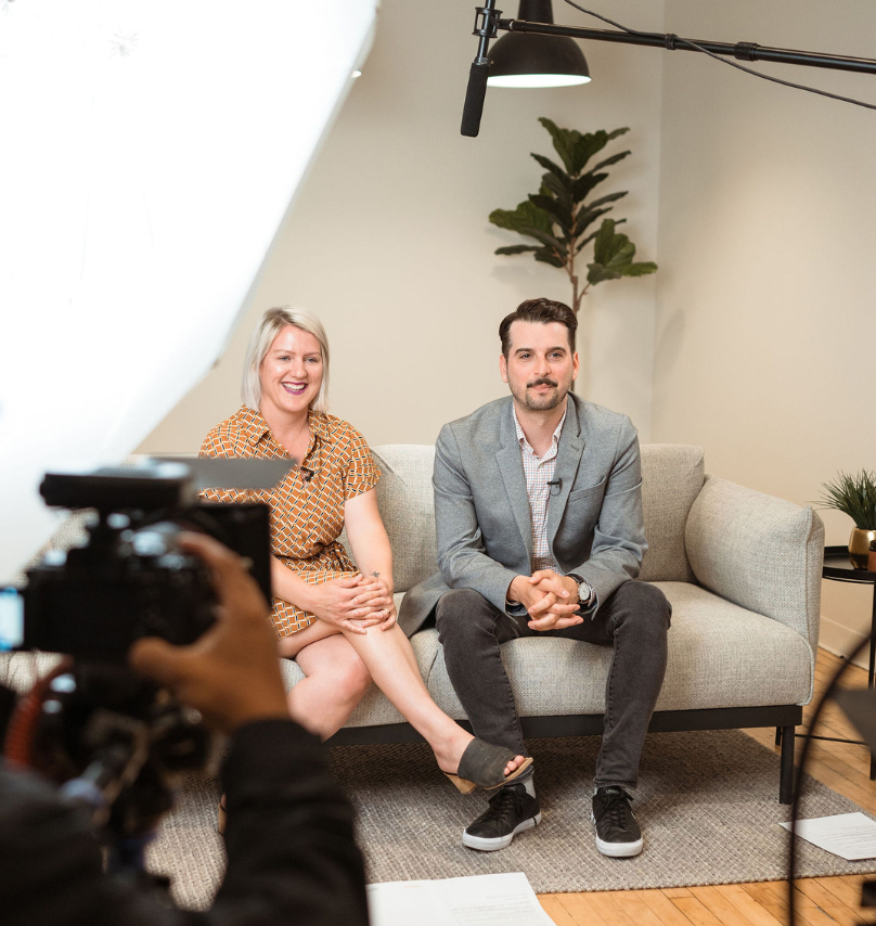 A behind-the-scenes photo of Matt and Brenlee filming a video while sitting on a couch. Dean is behind the camera adjusting camera settings.