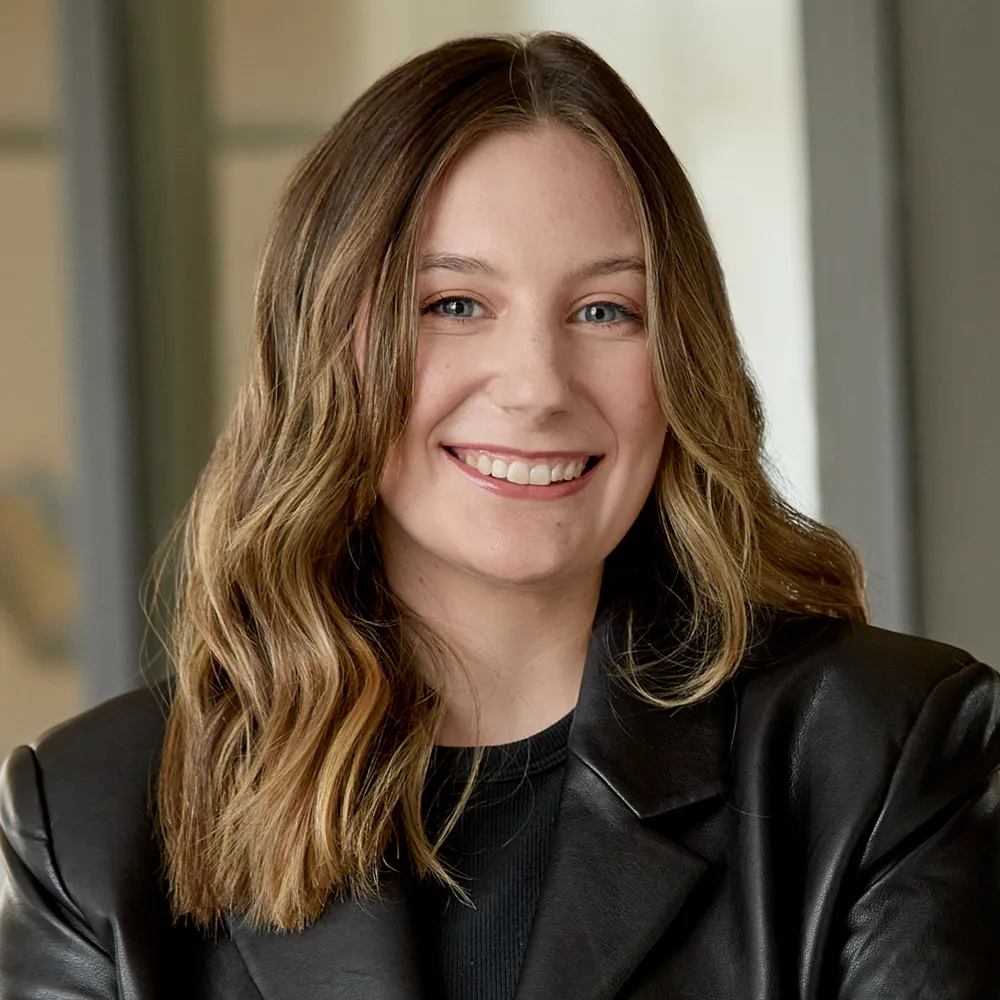 Headshot of Brigit Harvey. She is smiling, wearing a black shirt with a black leather jacket. She has long brunette hair with blonde highlights.