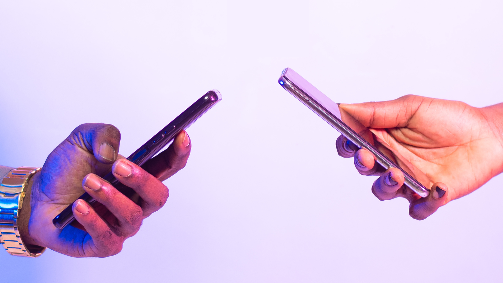 Two hands holding cell phones across from one another against a light purple backdrop. The hand on the left is wearing a gold watch. There is a purple hue to the photo.
