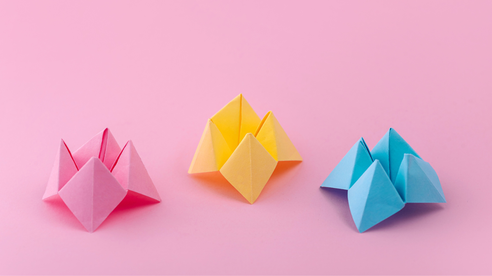 Three coloured paper fortune tellers (pink, yellow and blue), on a pink background.