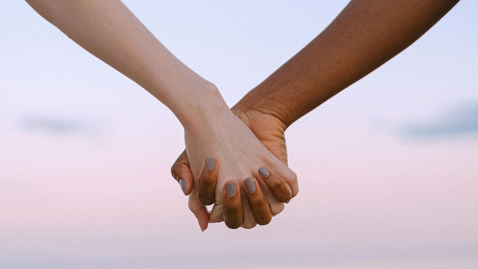 A close-up on two people's clasped hands with a pink sunset sky as the background.