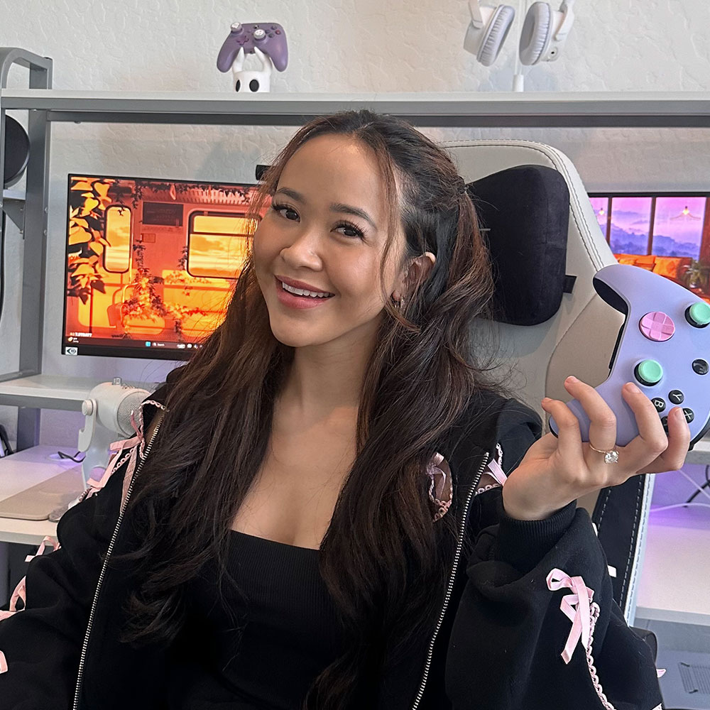 Jenna with long, wavy hair, smiling while holding a pastel purple gaming controller in her right hand. She is seated in front of a gaming setup, which includes a large monitor displaying a warm-toned game scene. She is wearing a black top with a black jacket that has pink ribbon details. The background shows additional gaming accessories, including a white and purple controller on a shelf.