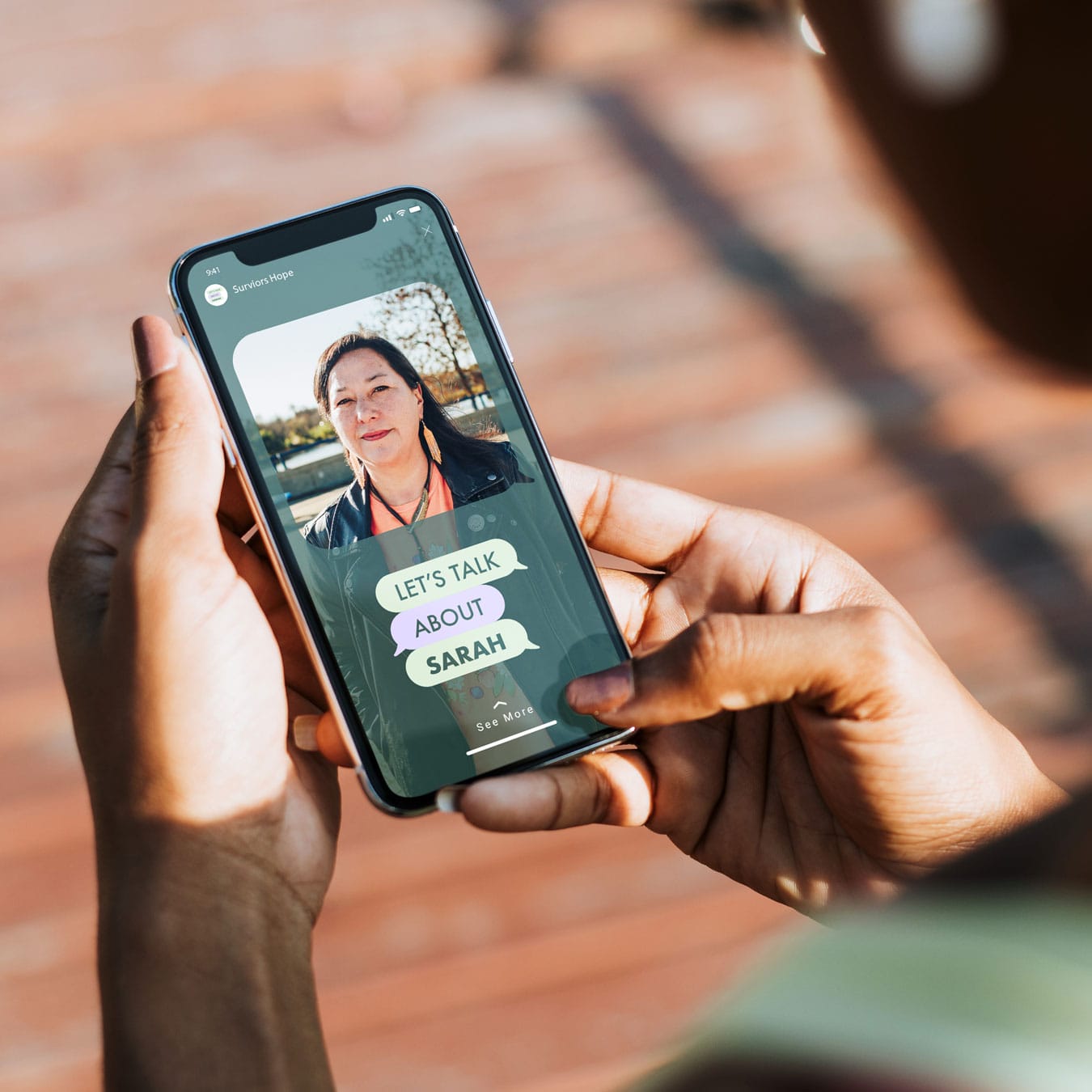 SARAH_Socials_5 The image shows a person holding a smartphone with an Instagram Story displayed on the screen. The story features a photo of a woman wearing a black jacket and orange top, with a tree-lined background. Overlay text on the story reads "LET'S TALK ABOUT SARAH" in colorful speech bubble graphics.