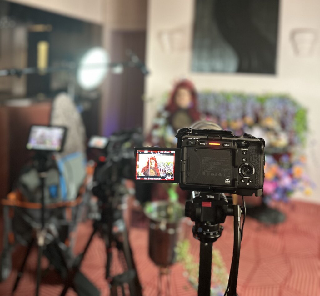 CGLCC Rose Bar Shoot A close-up image of a professional camera filming a person in front of a colourful floral backdrop, with lighting and video equipment blurred in the background.