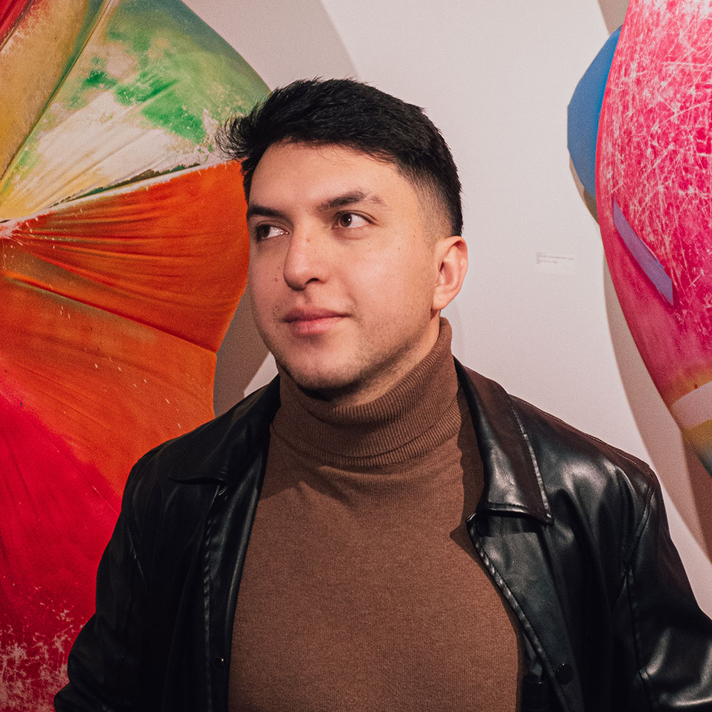 Juan standing confidently in front of a colorful abstract art piece, wearing a brown turtleneck and black leather jacket.