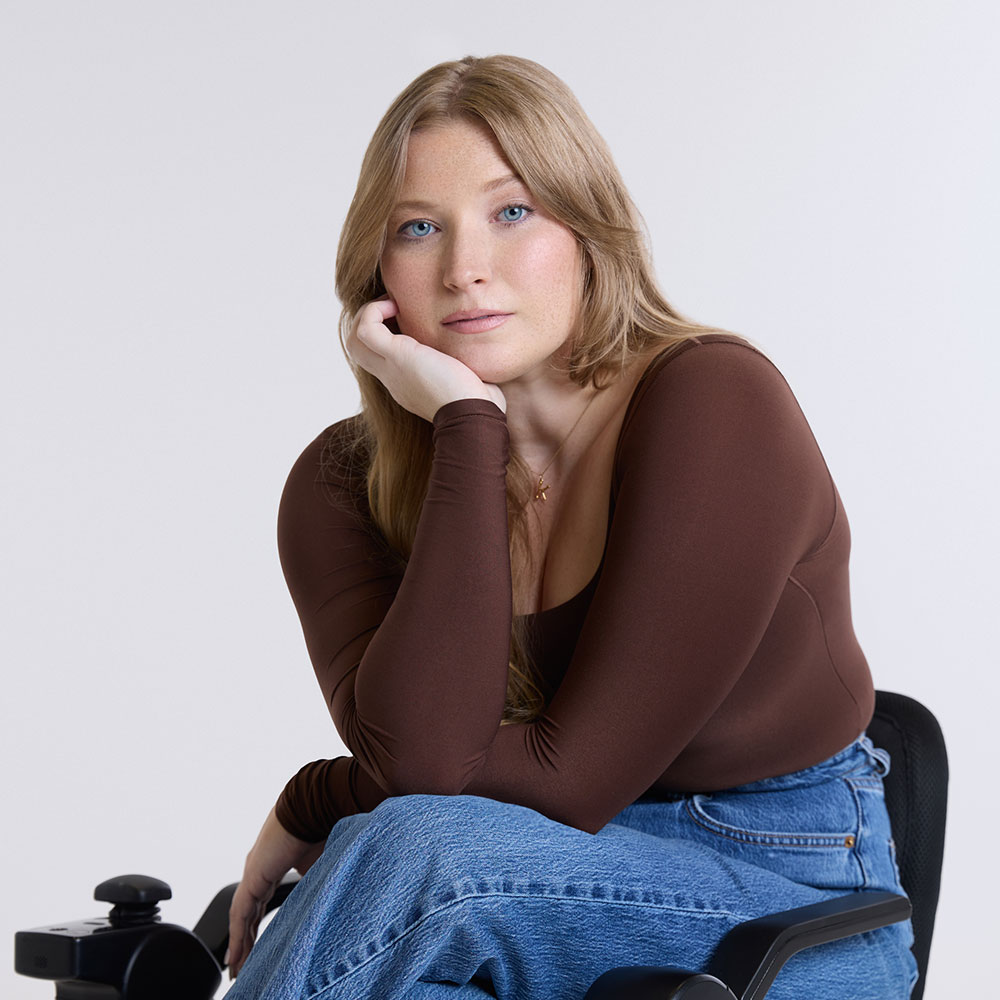 Kelcie, with long blonde hair, sitting thoughtfully in a wheelchair, wearing a brown long-sleeve top and blue jeans against a clean white background.