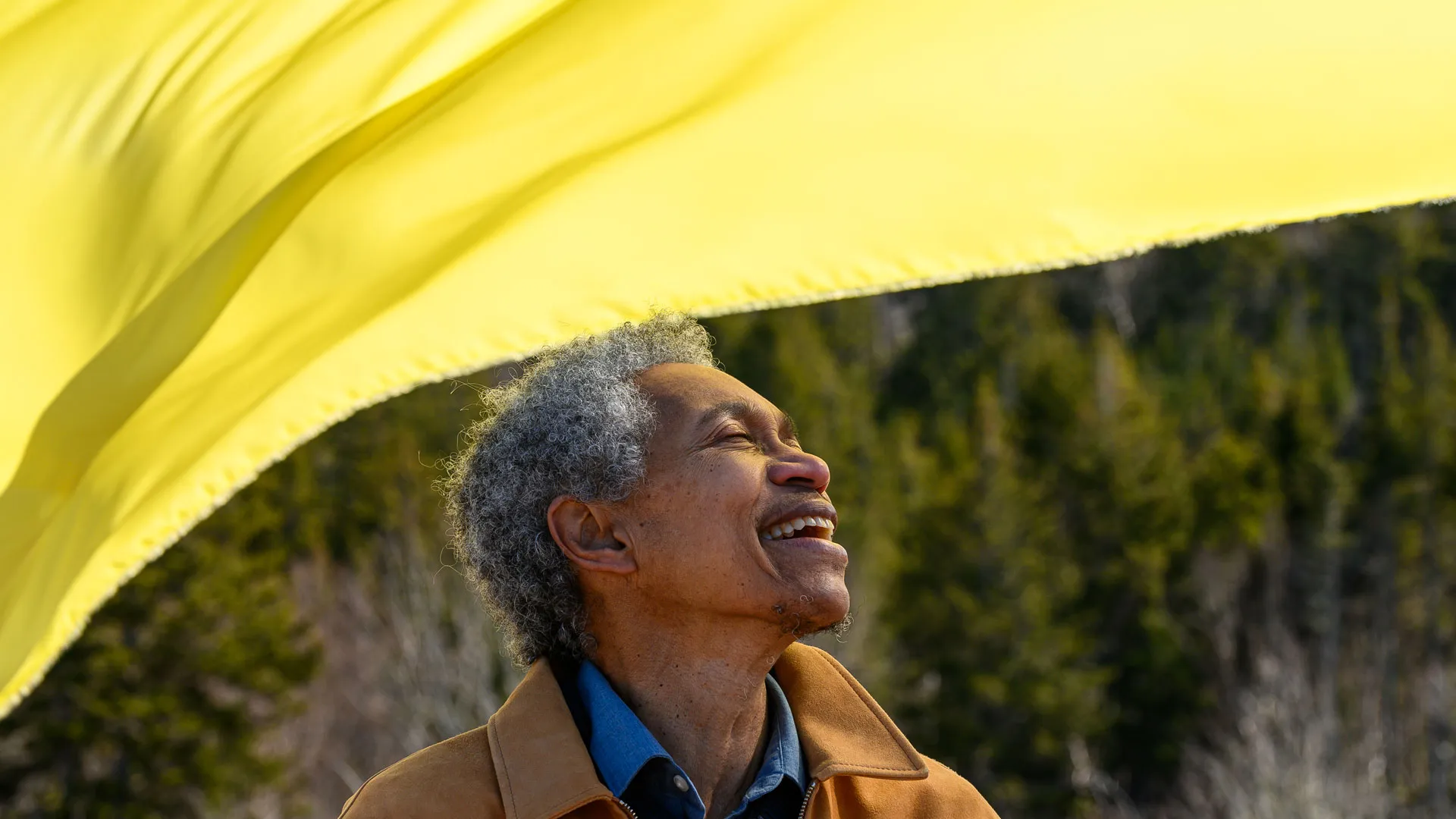 Beverly Glenn-Copeland with curly gray hair smiles with his eyes closed, basking in the sunlight. He wears a brown jacket over a denim shirt. A large yellow fabric billows in the wind above him.