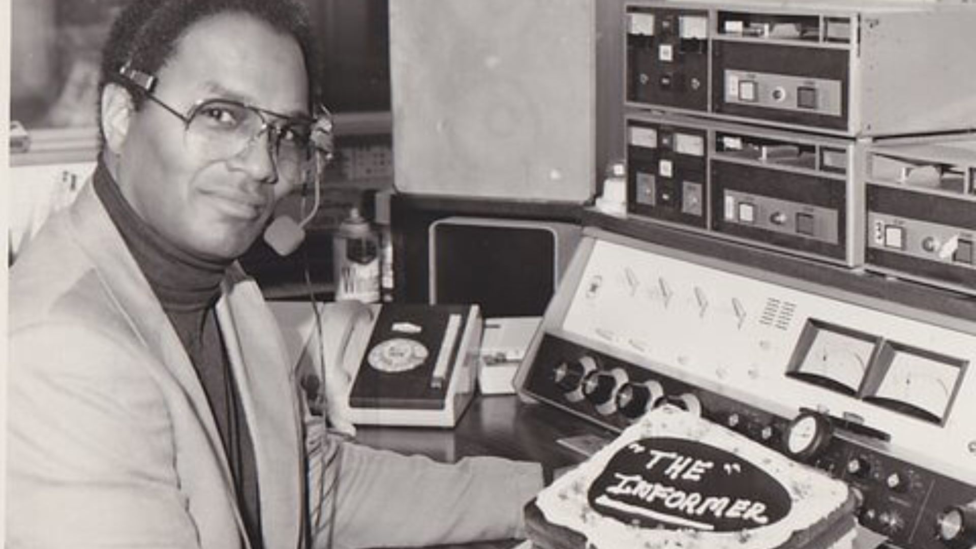 A black-and-white photograph of a Fil Fraser seated at a radio broadcasting station, wearing glasses and a headset with a microphone. He is dressed in a light-colored blazer over a dark turtleneck. In front of him on the control panel is a decorated cake with the words "The Informer" written on it. The background features vintage radio equipment, including tape decks and dials.