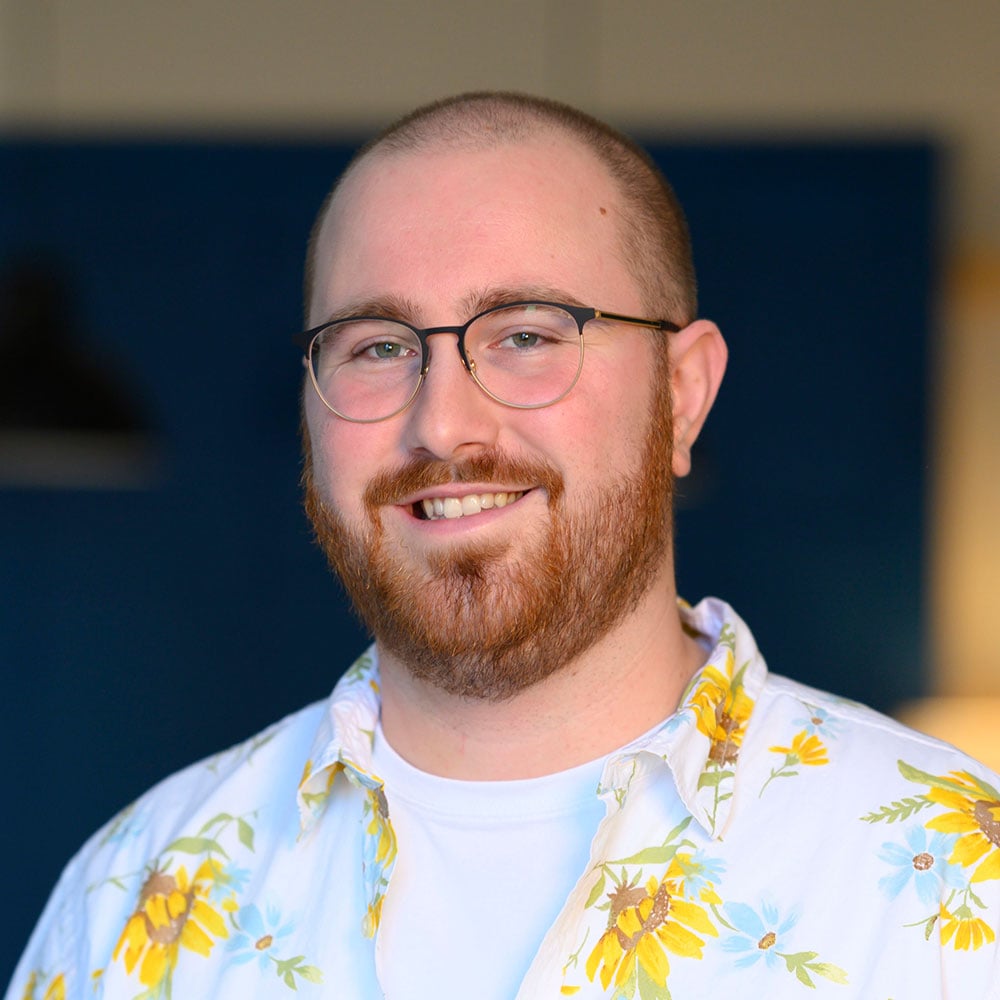Justin smiles warmly at the camera in a well-lit indoor setting. He has a neatly trimmed red beard, wears round glasses, and a white shirt with a vibrant yellow floral pattern over a plain white tee. The background is softly blurred with blue and neutral tones.