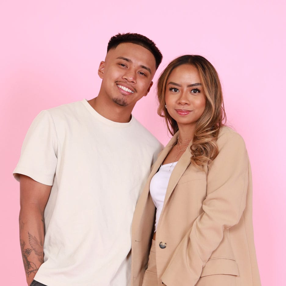 LoizzaRay-headshot : Loizza and Ray standing together in front of a pink background. Ray is wearing a cream-colored t-shirt and smiling with his arm gently behind Loizza. Loizza is wearing a beige blazer over a white top, with her hair styled in loose curls, smiling softly at the camera.