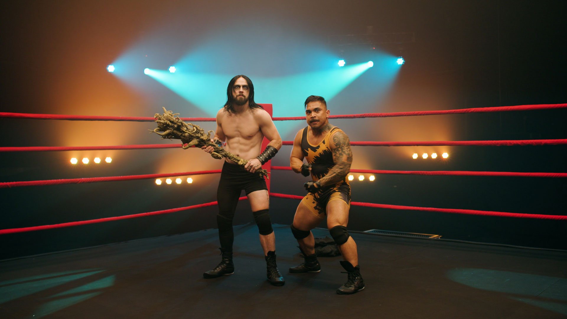 BY-TruFlex-002 Two wrestlers pose in a dramatic, smoke-filled wrestling ring under colorful stage lights. One wears black shorts and boots, holding a large, twisted tree branch prop. The other, in a gold and black singlet with tattoos on his arm, crouches in a ready stance. This is a still from the BrettYoung TruFlex campaign video.