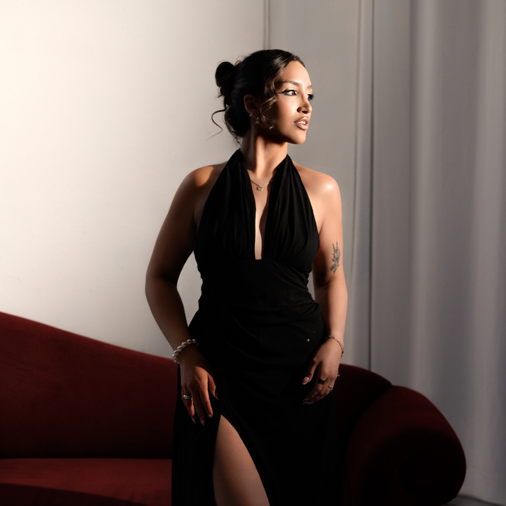 Luciana wearing a black halter-style dress with a thigh-high slit stands beside a deep red curved sofa, looking toward natural light streaming in from the right. Her dark hair is styled in a sleek bun, and she wears delicate jewelry and subtle makeup. The portrait has a warm, elegant feel with soft shadows and a minimalist white backdrop.