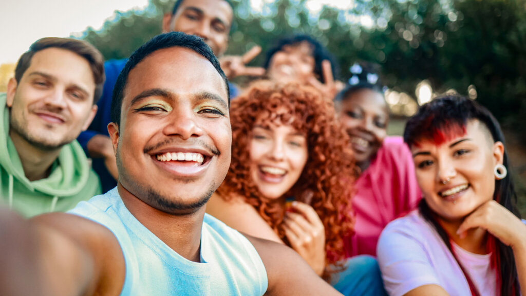 Young diverse people smiling at mobile phone camera at city park. African nonbinary taking selfie with friends. LGBT community Gen Z pride goers