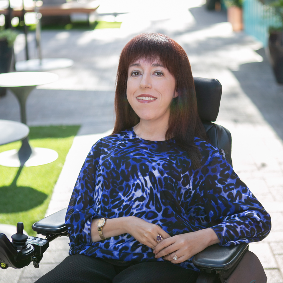 Jamie smiling with shoulder-length dark auburn hair sits in a power wheelchair outdoors on a sunny day. She is wearing a blue patterned long-sleeve top, black pants, and simple jewelry, with her hands resting comfortably in her lap. The background features a bright patio with tables, greenery, and soft natural light.